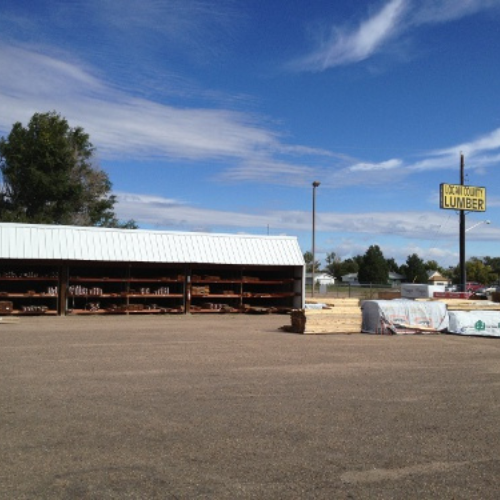 A large outdoor lumber yard under a partly cloudy sky, with stacks of wood and a sign that reads 