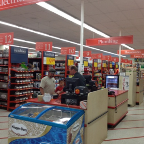A man and child stand at a checkout counter in a store with red aisle signs. The atmosphere is busy and organized, with shelves filled with products.