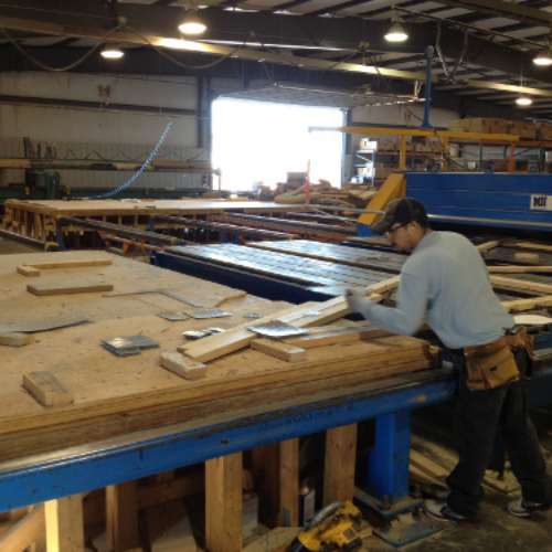 A worker in a cap and gloves is assembling wooden panels on a large table inside a factory. The scene is busy, with tools and materials around.