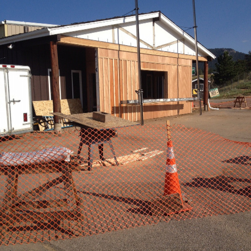 Construction scene with a partly built wooden structure, orange safety netting, and traffic cones. A person is sawing wood in sunlight.