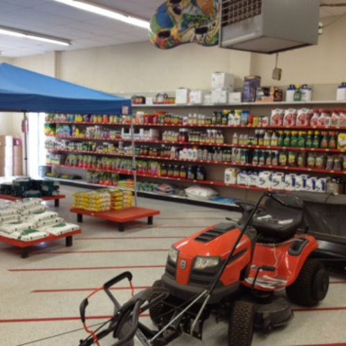 A store interior with shelves stocked with gardening supplies. Foreground shows a red lawnmower and a blue canopy. Atmosphere is organized and practical.