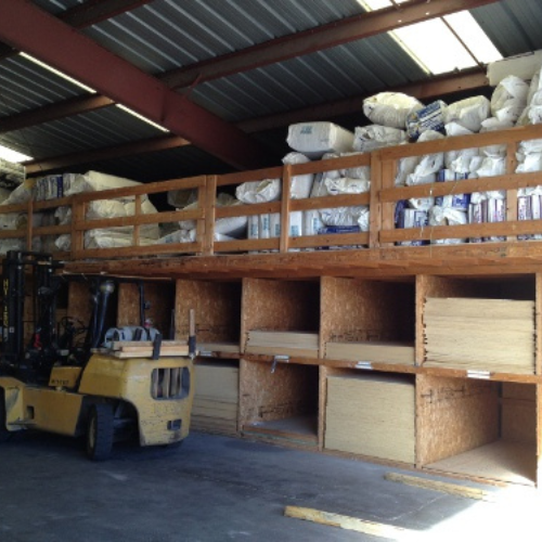 A warehouse interior with a forklift near stacked plywood sheets. Bags of insulation are stored on a wooden loft above, under a metal roof.