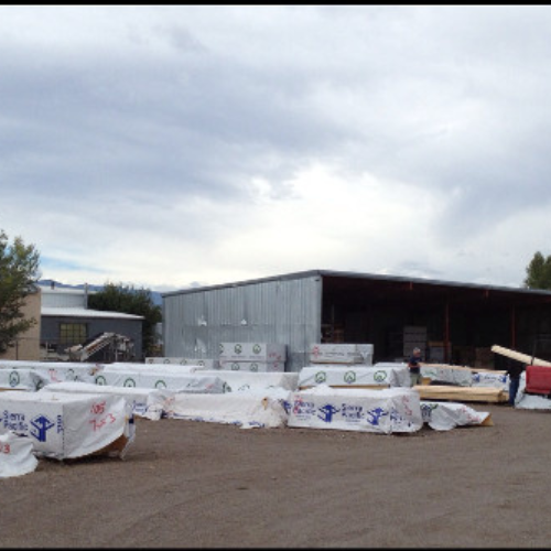 Outdoor lumber yard with stacked, wrapped wood piles near a large metal warehouse under a cloudy sky. Workers are visible, indicating activity.