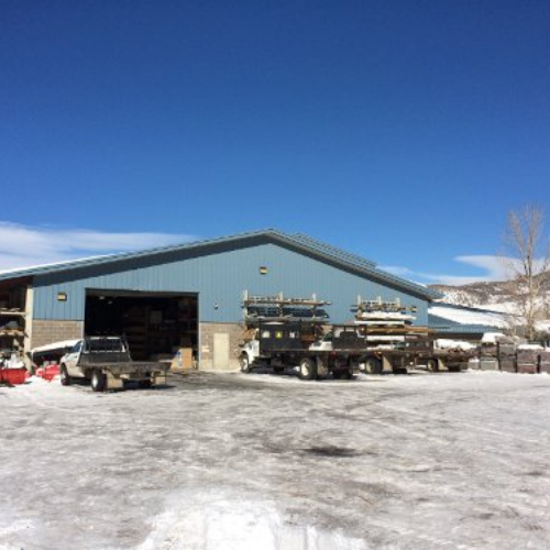 A warehouse with a blue metal roof is surrounded by snow and vehicles loaded with supplies. The clear blue sky adds a crisp, cold ambiance.