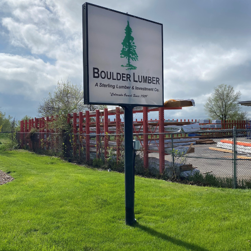A sign for Boulder Lumber on a lush green lawn under a cloudy sky, with piles of wood and red fencing visible in the background.