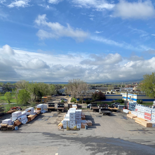 Industrial storage yard with stacks of lumber and construction materials under a partly cloudy sky. Green fields and distant trees surround the area.