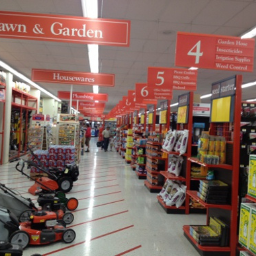 Wide aisle in a hardware store with lawn mowers on the left, shelves of garden supplies on the right, and overhead signs indicating departments.
