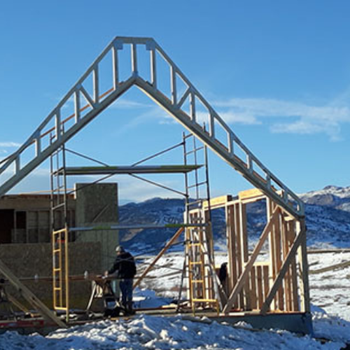 A construction site with workers assembling the frame of a building against a backdrop of mountains and a clear blue sky.
