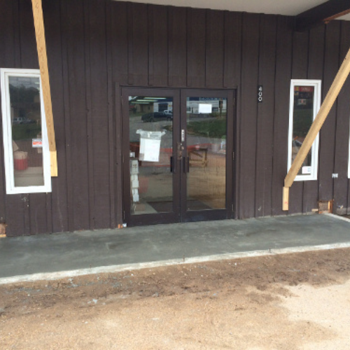A brown wooden building facade with glass double doors in the center, flanked by two tall narrow windows, under construction with wood supports.