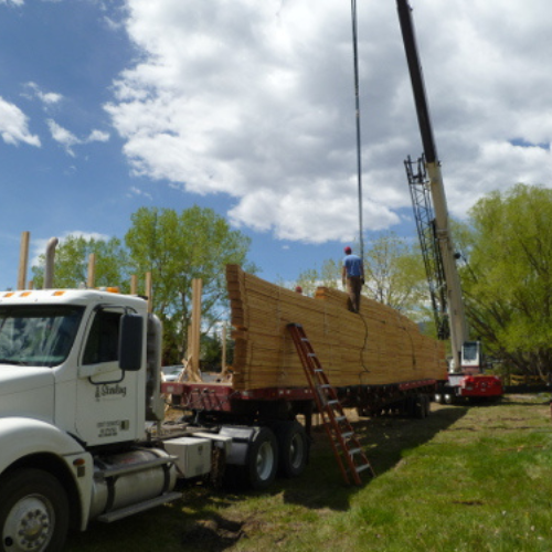 A crane lifts a large stack of lumber from a flatbed truck under a partly cloudy sky. A worker stands on top, wearing protective gear. Greenery surrounds the scene.
