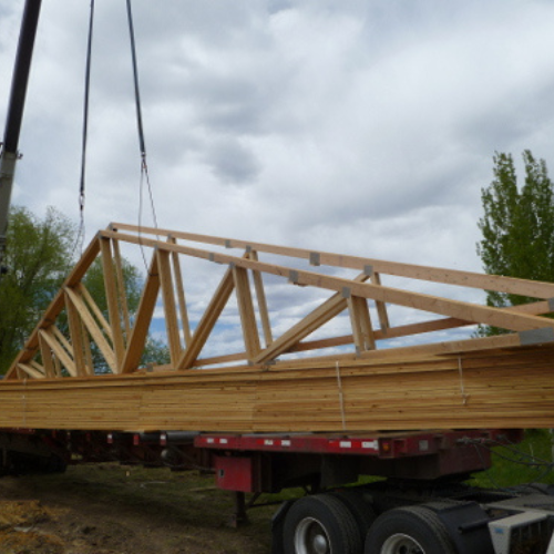 A large wooden truss is being lifted by a crane from a flatbed truck on a construction site. Cloudy sky and green trees are in the background.