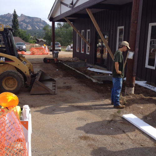 A construction worker stands near a brown building with leaning timber supports. A skid steer loader is parked nearby. Orange safety netting surrounds the site.