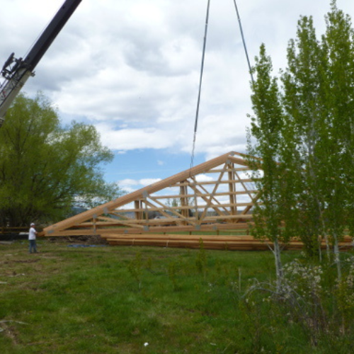 A crane lifts a large wooden roof truss on a grassy field, surrounded by trees under a cloudy sky. A person stands nearby, observing.