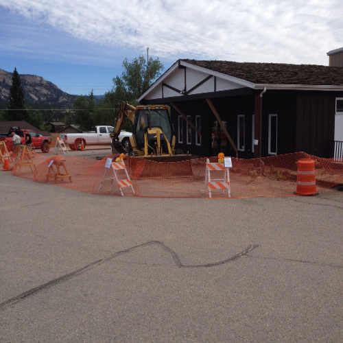 Small construction site outside a dark brown building, surrounded by orange netting and traffic cones. A backhoe is parked nearby under a cloudy sky.