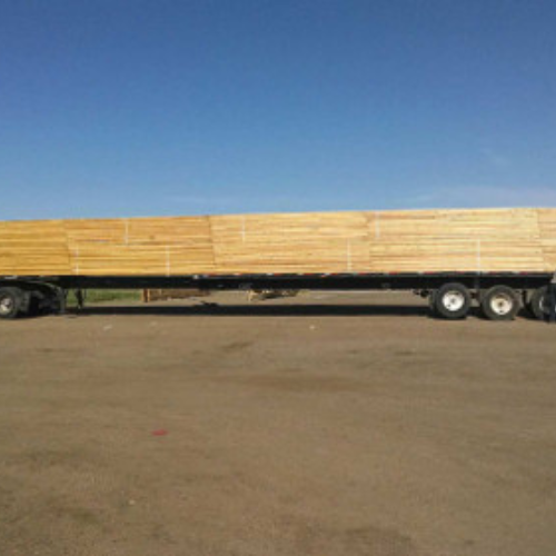 A flatbed truck loaded with stacked lumber, parked on a paved surface under a clear blue sky, conveying a sense of industry and transportation.