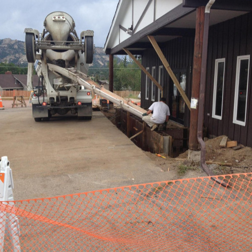 A concrete mixer truck pours cement alongside a building, with wooden supports bracing it. A worker sits nearby, overseeing the process. The sky is overcast.