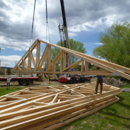 Large wooden trusses are being lifted by a crane at a construction site under a cloudy sky, with workers and equipment visible, creating a busy atmosphere.