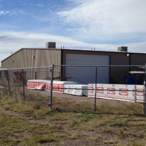 Industrial building behind a chain-link fence, with stacked construction materials on grass. Overcast sky suggests a quiet, utilitarian setting.