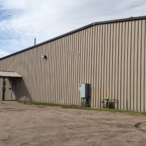 Large industrial warehouse with beige metal siding, small entrance, utility boxes, and a gravel lot. Overcast sky conveying an industrial tone.
