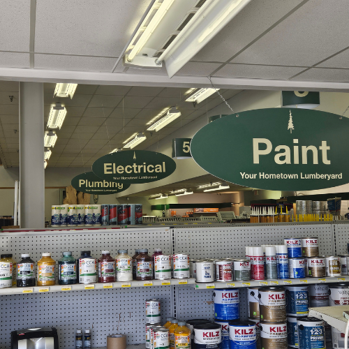 Hardware store interior with shelves of paint cans in the foreground. Overhead signs label sections like 