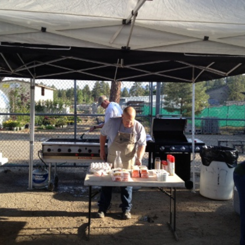 A man stands under a canopy at an outdoor barbecue event, organizing food on a table. Grills are in the background, with trees and a fenced area nearby.