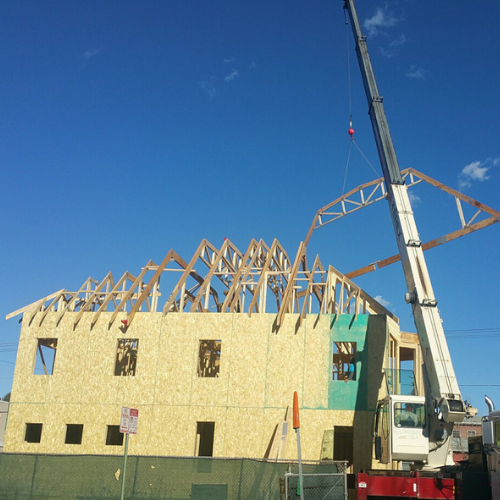 A crane lifts a wooden truss onto a two-story building under construction against a clear blue sky, conveying progress in the construction process.