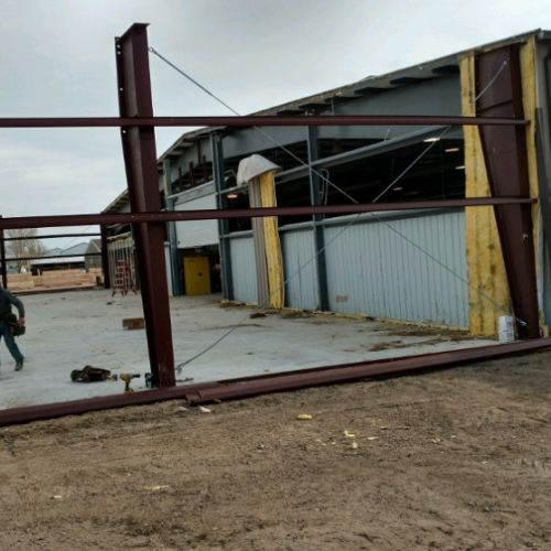 A partially constructed metal building stands on a dirt lot under a cloudy sky. Steel beams and exposed insulation are visible, with a worker nearby.