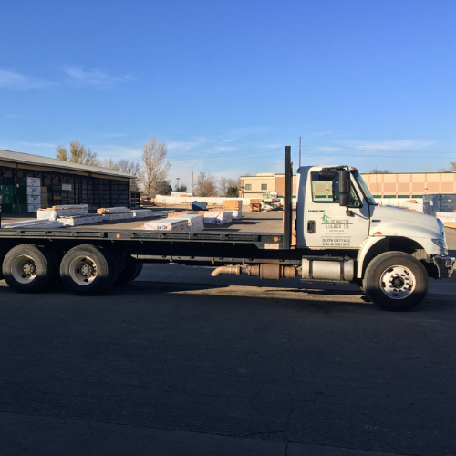 A white flatbed truck laden with construction materials is parked in an industrial area under a clear blue sky, evoking a sense of productivity.