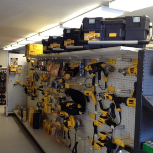 A store aisle displays various yellow and black power tools on a pegboard, including drills and saws. A black tool case is on the top shelf.