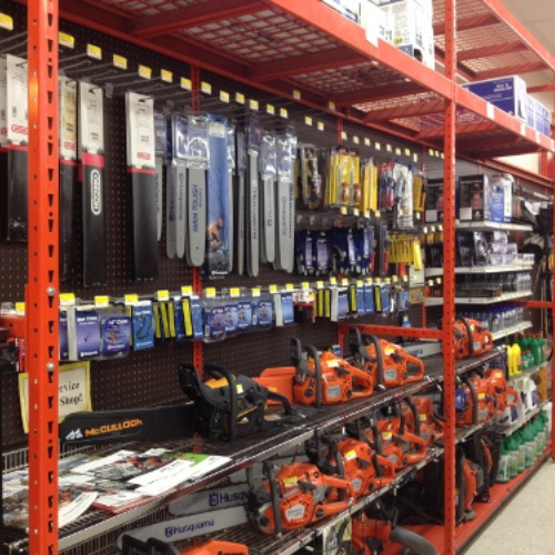 A store aisle with orange chainsaws, packaged chainsaw chains, and assorted hardware on shelves. Tools are neatly arranged, creating an organized display.