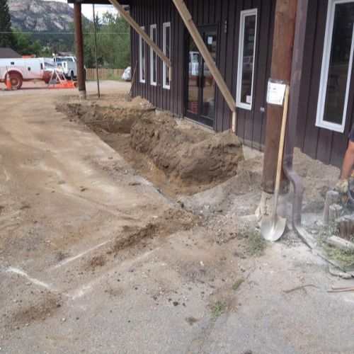 Dirt excavation by a building with large windows, wood beams, and a shovel nearby. A construction vehicle is visible in the distance on a cloudy day.