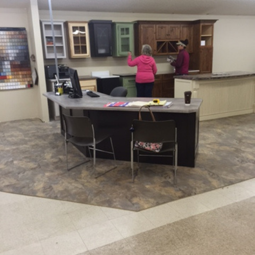 A showroom features two women discussing kitchen cabinets on display. A counter with chairs is in the foreground, setting a professional tone.