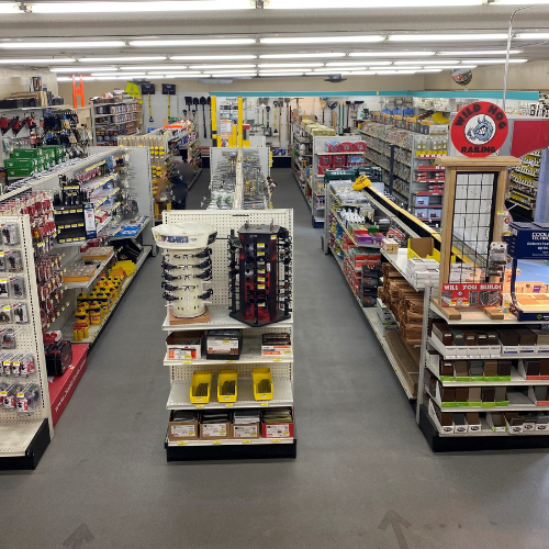 A well-lit hardware store interior with neatly arranged aisles filled with tools, paint cans, and accessories. The space conveys order and variety.