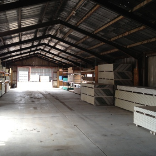 A spacious, empty warehouse with a metal roof, stacked with organized piles of wooden and drywall sheets along the sides, lit by natural light.