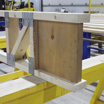A wooden structure being assembled, featuring a rectangular piece of wood joined by metal brackets to a frame, on a yellow workbench.