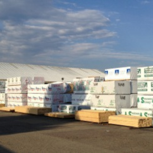 Stacks of wrapped lumber are neatly arranged outdoors on asphalt, with a large building in the background under a partly cloudy sky.