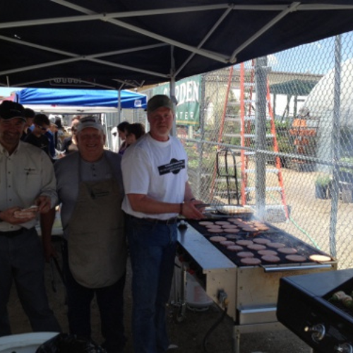 Three men cooking burgers on a large grill under a canopy at an outdoor event. They smile at the camera, creating a friendly, community atmosphere.