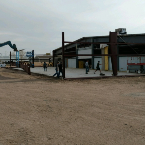 Construction workers assemble a steel framework for a new building extension on an overcast day. Machinery and tools are visible in the background.