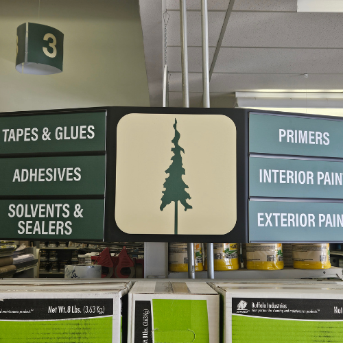 Store aisle sign with a tree symbol, labeled for tapes, adhesives, primers, and paints. Shelves hold various paint cans beneath the sign.