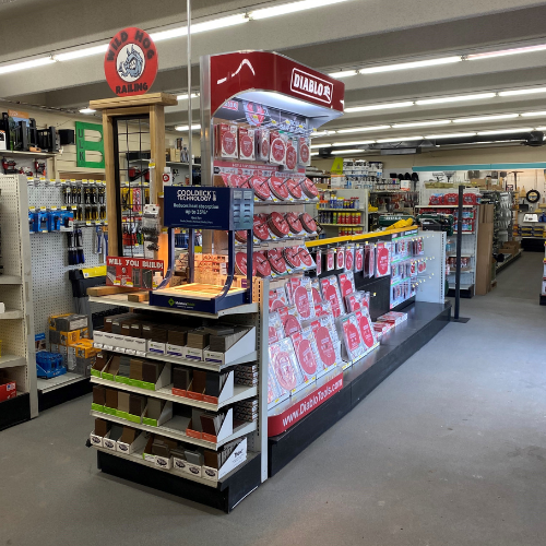 A hardware store aisle featuring a Diablo display stand with various circular saw blades. Shelves are filled with tools and supplies under bright lighting.