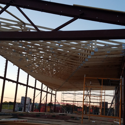 Construction site at sunset with a partially built wooden roof supported by steel beams. Scaffolding is visible, suggesting active progress.
