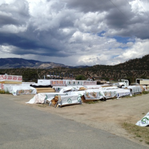 Outdoor lumber yard with stacks of covered wood, cloudy sky, and distant mountains. The setting is calm, with a mix of natural and industrial elements.