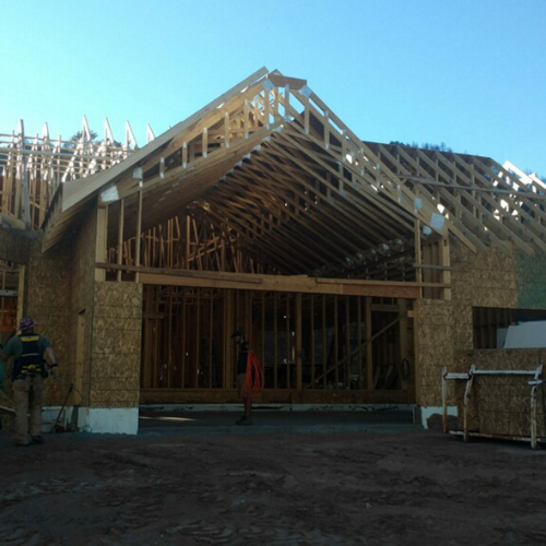 A wooden house frame under construction with exposed beams and trusses. Workers are present, and the ground is covered in construction materials.