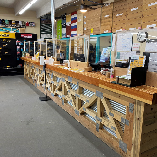 A hardware store checkout area with a long wooden counter, clear protective screens, and organized tool displays. The atmosphere is practical and orderly.