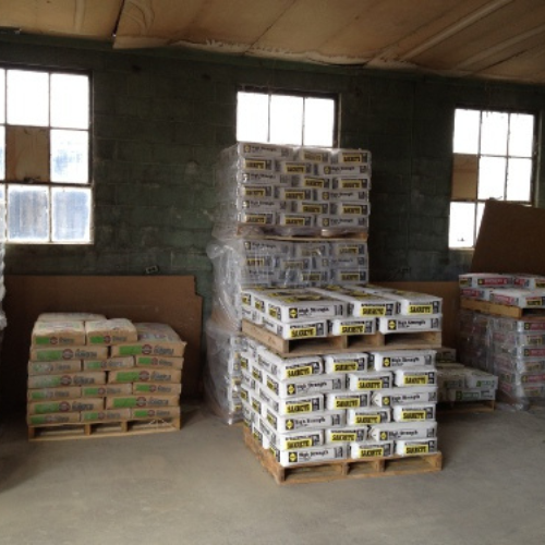 Stacks of packaged building materials on wooden pallets in a dimly lit warehouse. Three large windows illuminate the gray brick walls.