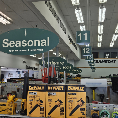 Decorated aisle in a hardware store with seasonal signs and tools. Prominent DeWalt boxes in the foreground. Bright overhead lighting.