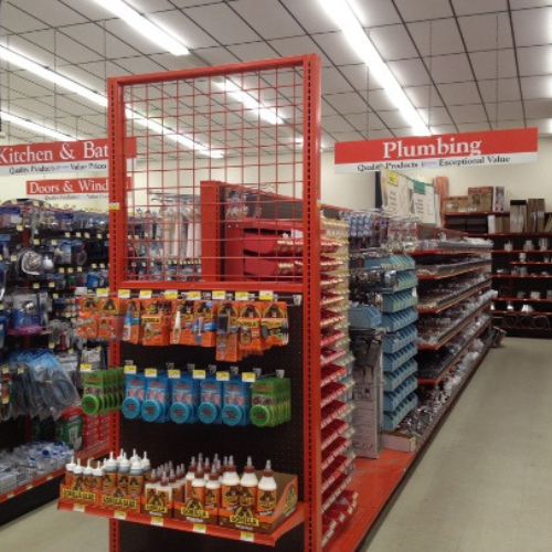 A hardware store aisle with shelves of plumbing supplies and adhesives. Overhead signs read 