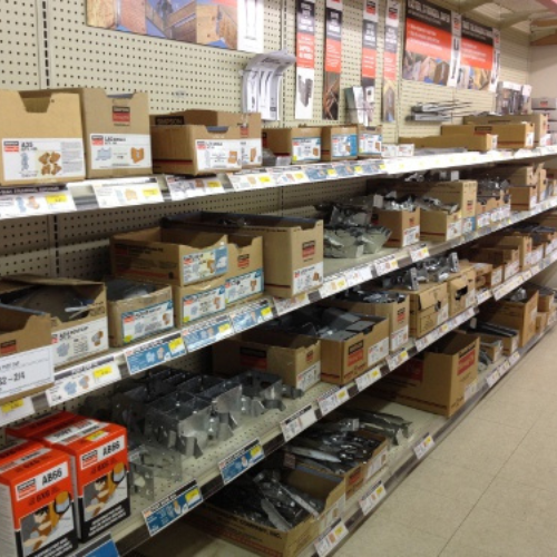 A hardware store aisle with organized shelves displaying boxes of metal brackets and fasteners. Labels and prices are visible, creating a tidy, utilitarian feel.