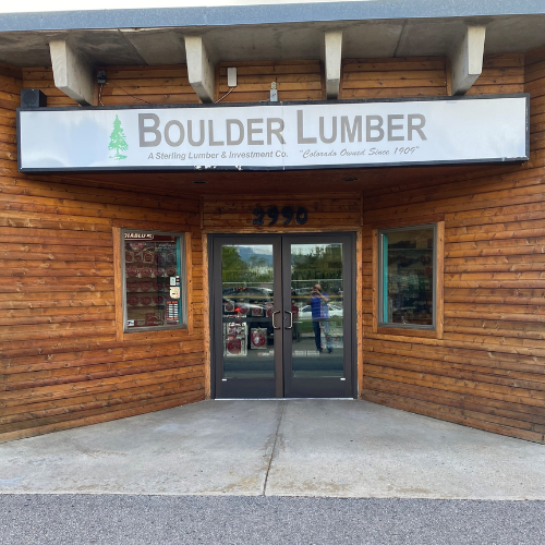 Storefront of Boulder Lumber, featuring wooden paneling, a white sign with green tree logo, and glass double doors reflecting the outdoor surroundings.