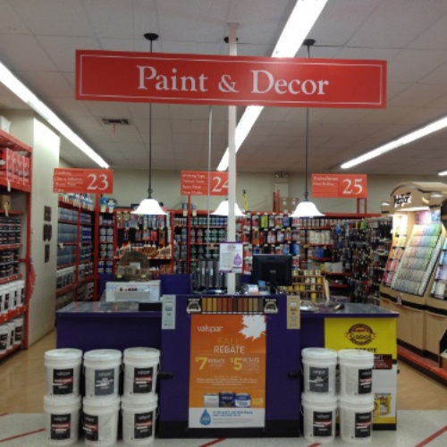 A hardware store's paint and decor section with rows of paint cans, colorful paint swatches, and rebate signs. A service counter is at the center.